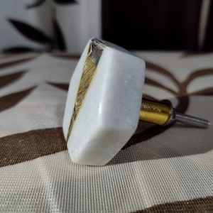 Pode incluir: Um puxador de arm&aacute;rio em m&aacute;rmore branco com um detalhe dourado. O puxador tem formato quadrado com bordas chanfradas e uma haste de metal dourado. A ferragem &eacute; projetada para uso em gavetas, arm&aacute;rios ou m&oacute;veis.