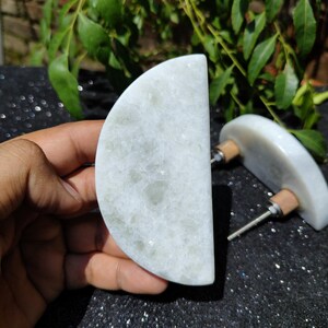 May include: Two white marble cabinet knobs with a semi-circular design. Each knob has a wooden base and a metal screw. The knobs are displayed against a dark, speckled surface with green foliage in the background.