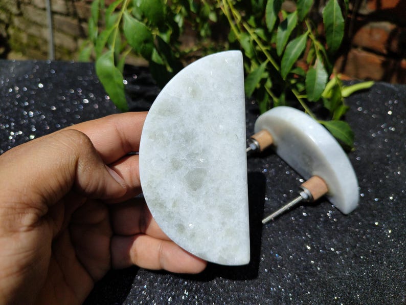 May include: Two white marble cabinet knobs with a semi-circular design. Each knob has a wooden base and a metal screw. The knobs are displayed against a dark, speckled surface with green foliage in the background.