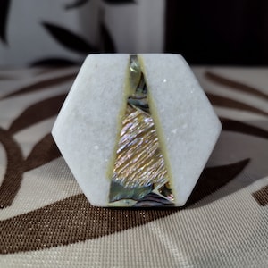 May include: A white marble hexagonal decorative object with a central inlay of iridescent abalone shell and gold-toned material. The object has a polished finish and is likely a decorative element or a small sculpture.