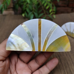 May include: A decorative, semi-circular object with a white, mother-of-pearl surface, accented with gold and iridescent stripes. The object is likely a decorative item or a small dish, with a second one visible in the background.