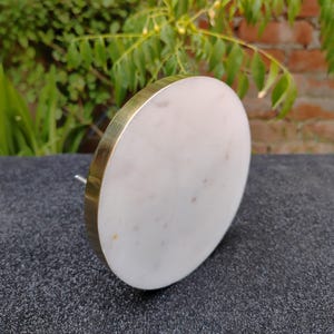 May include: A white marble cabinet knob with a gold-colored metal rim. The knob is round and has a silver-colored screw on the back. The background is a dark gray surface with green foliage and a brick wall.