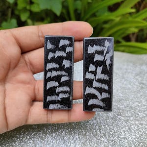 Op de afbeelding: Twee rechthoekige zwarte oorbellen met een veervormig patroon in grijstinten. De oorbellen worden in een hand gehouden tegen een vage groene en zilveren achtergrond. De oorbellen zijn waarschijnlijk voor doorboorde oren.