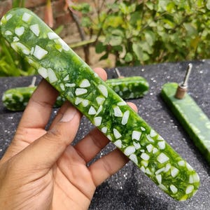 Puede incluir: Un objeto rectangular verde y blanco con un patrón moteado se sostiene en una mano. El objeto es de color verde claro con formas blancas irregulares. Otros objetos similares están en el fondo.