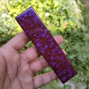 Op de afbeelding: Een rechthoekig, handgemaakt object met een diepe bordeauxrode basis en wervelende paarse accenten. Het object wordt in een hand gehouden tegen een vage groene achtergrond, wat de gepolijste, glanzende afwerking benadrukt. Het lijkt een decoratief element te zijn.