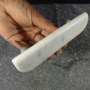 May include: A white marble cabinet pull with a rectangular shape and rounded edges. The pull has a silver-coloured screw and a small, round, grey-coloured base. The marble has subtle grey veining.