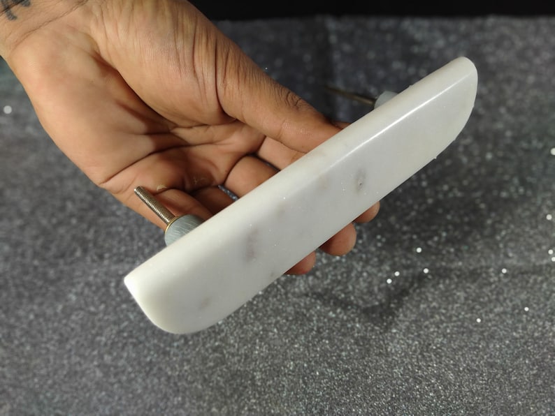 May include: A white marble cabinet pull with a rectangular shape and rounded edges. The pull has a silver-coloured screw and a small, round, grey-coloured base. The marble has subtle grey veining.