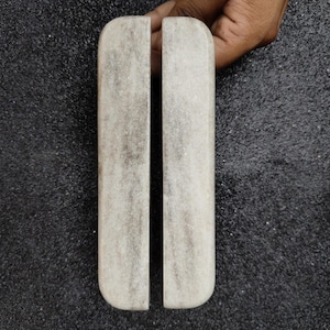 Puede incluir: Dos piezas rectangulares de mármol claro con bordes redondeados, posiblemente manijas o elementos decorativos. El mármol tiene un patrón veteado natural. Los objetos están sobre un fondo oscuro y texturizado.