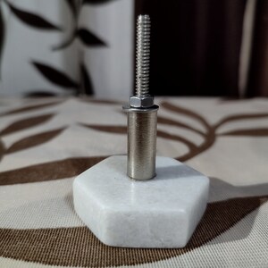 Pode incluir: Puxador de arm&aacute;rio hexagonal em m&aacute;rmore branco com um pino, parafuso e arruela de metal prateado. O puxador &eacute; projetado para m&oacute;veis e tem uma est&eacute;tica moderna.