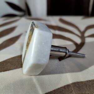 Pode incluir: Um puxador de arm&aacute;rio de m&aacute;rmore branco com formato quadrado e um detalhe dourado. O puxador possui um parafuso de metal prateado para fixa&ccedil;&atilde;o. O fundo &eacute; um tecido estampado.