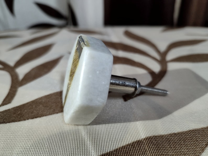 Pode incluir: Um puxador de arm&aacute;rio de m&aacute;rmore branco com formato quadrado e um detalhe dourado. O puxador possui um parafuso de metal prateado para fixa&ccedil;&atilde;o. O fundo &eacute; um tecido estampado.