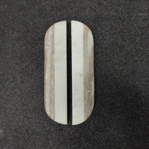 May include: Two matching, elongated, oval-shaped stones, each bisected by a vertical line. The stones have a light, off-white color with subtle gray veining, set against a dark gray background. The stones appear to be polished and smooth.