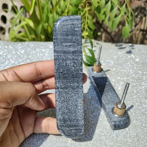May include: Two grey stone cabinet pulls with a textured surface. Each pull has a rectangular shape with rounded edges and a metal screw for installation. The pulls are displayed on a reflective surface with greenery in the background.