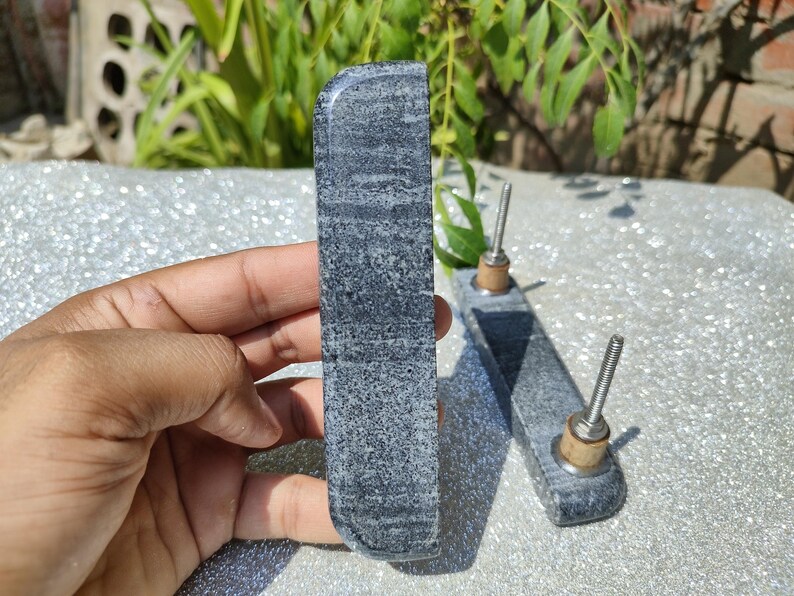 May include: Two grey stone cabinet pulls with a textured surface. Each pull has a rectangular shape with rounded edges and a metal screw for installation. The pulls are displayed on a reflective surface with greenery in the background.