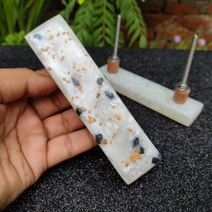 Könnte beinhalten: Zwei weiße Schubladengriffe aus Harz mit schwarzen und goldenen Flecken. Die Griffe sind rechteckig und haben eine glatte, polierte Oberfläche.