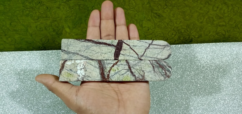 May include: Two rectangular, polished stone pieces with rounded ends. The stone has a light green base colour with dark brown veining throughout. The pieces are held in a hand against a green and silver background.