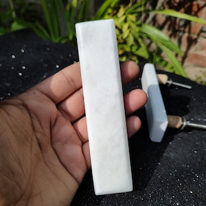 Puede incluir: Dos tiradores de cajón rectangulares de mármol blanco. El tirador más grande se sostiene en una mano. El tirador más pequeño tiene dos agujeros para el montaje.