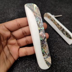 Pode incluir: Dois botões de armário brancos com incrustações de madrepérola de abalone iridescente. Os botões têm a forma de um retângulo arredondado com uma ligeira curva.