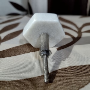 Pode incluir: Um puxador de arm&aacute;rio de m&aacute;rmore branco com formato hexagonal. O puxador possui uma haste roscada prateada com porca e arruela. O puxador &eacute; projetado para uso em arm&aacute;rios e gavetas.