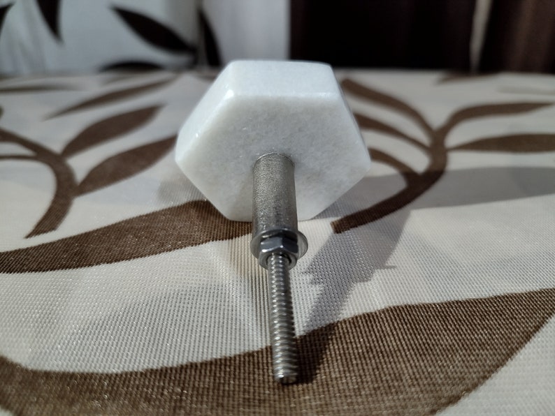 Pode incluir: Um puxador de arm&aacute;rio de m&aacute;rmore branco com formato hexagonal. O puxador possui uma haste roscada prateada com porca e arruela. O puxador &eacute; projetado para uso em arm&aacute;rios e gavetas.