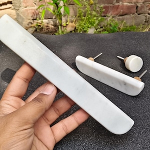 Op de afbeelding: Witte marmeren kastbeslagset, bestaande uit een lange rechthoekige handgreep en twee kleinere stukken met metalen pinnen. Het beslag heeft een gladde, gepolijste afwerking en een natuurlijk adermotief. Voor kasten en lades.