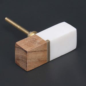 May include: A rectangular cabinet knob with a wood and white marble design. The knob has a brass screw and a small brass accent. The wood is light brown, and the marble is white.