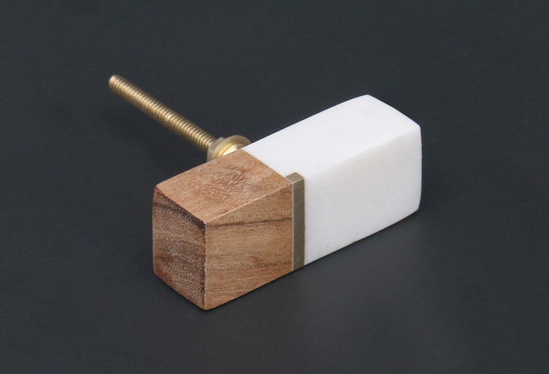 May include: A rectangular cabinet knob with a wood and white marble design. The knob has a brass screw and a small brass accent. The wood is light brown, and the marble is white.