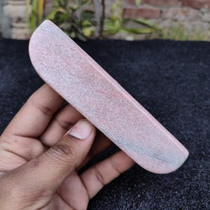 May include: A pink and grey stone object with a curved, rectangular shape. The stone has a speckled texture and is held in a hand. The object appears to be a sharpening stone or similar tool.