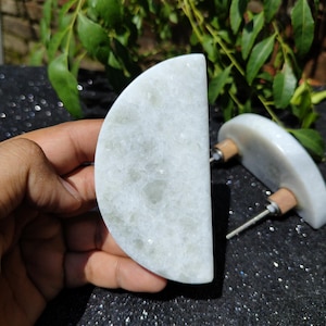 May include: Two white marble cabinet knobs with a semi-circular design. Each knob has a wooden base and a metal screw. The knobs are displayed on a dark, glittery surface with green foliage in the background.