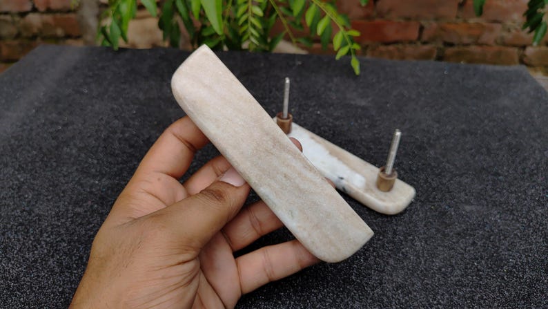 May include: Two light-coloured, rectangular cabinet pulls with metal posts and brass-coloured accents. One pull is held in a hand, while the other rests on a dark, textured surface. The pulls have a smooth, curved design.