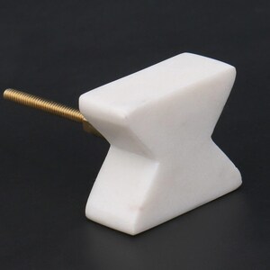 May include: A white marble cabinet knob with a brass screw. The knob has an X-shaped design. The hardware is set against a black background.