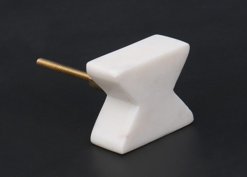 May include: A white marble cabinet knob with a brass screw. The knob has an X-shaped design. The hardware is set against a black background.