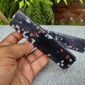 Könnte beinhalten: Zwei schwarze, rechteckige Harzrohlinge mit weißen und roten Flecken. Die Rohlinge werden in einer Hand gehalten.