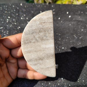 May include: A semi-circular, light grey marble stone with darker grey veining. The stone is held in a hand, highlighting its smooth, polished surface. The object is likely a decorative piece or a small functional item.