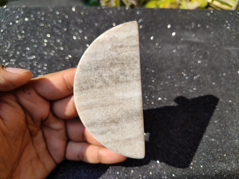 May include: A semi-circular, light grey marble stone with darker grey veining. The stone is held in a hand, highlighting its smooth, polished surface. The object is likely a decorative piece or a small functional item.