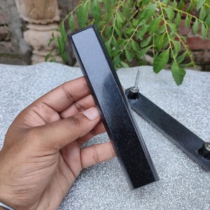 May include: Two black rectangular cabinet pulls with silver hardware. One pull is held in a hand, while the other lies on a textured surface. The pulls have a polished, reflective surface, and are likely made of stone or a similar material.