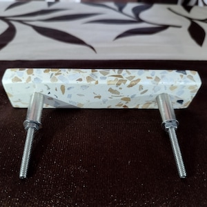 May include: A white, rectangular cabinet pull with a speckled terrazzo pattern. The pull has two silver screws.