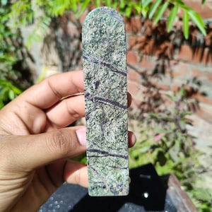 Op de afbeelding: Een rechthoekige, gepolijste steen met een afgeronde bovenkant. De steen heeft een groen en grijs gemarmerd patroon met donkergrijze aderen. Het object wordt in een hand gehouden, met een vage achtergrond van groen en een bakstenen muur.