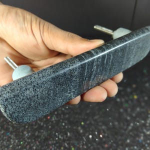 May include: A dark grey, rectangular stone door pull with two silver-coloured posts and light grey knobs. The stone has a speckled pattern and is held in a hand.