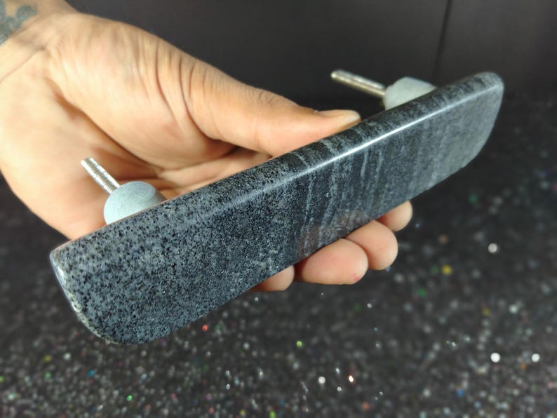May include: A dark grey, rectangular stone door pull with two silver-coloured posts and light grey knobs. The stone has a speckled pattern and is held in a hand.