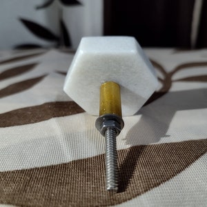 Pode incluir: Puxador de arm&aacute;rio hexagonal branco em m&aacute;rmore com haste dourada e parafuso prateado. O puxador &eacute; projetado para m&oacute;veis e tem uma est&eacute;tica moderna e minimalista. O hardware &eacute; adequado para gavetas e portas.