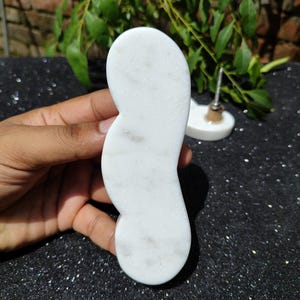 May include: A white, kidney-shaped marble gua sha tool. The tool is held in a hand against a black, glittery surface. A small, white, round object with a metal post is in the background.