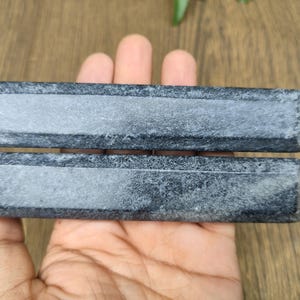 May include: Two rectangular, dark gray stone blocks with a lighter gray recessed center are held in a hand. The blocks have a textured surface and are likely used for sharpening or honing tools. The background is a wooden surface.