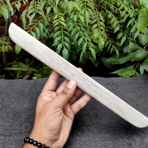 May include: A long, slender, off-white selenite wand with a smooth, polished surface and a slightly curved, elongated shape. The crystal displays a subtle, fibrous texture with natural variations in its pale, translucent, and opaque white color.