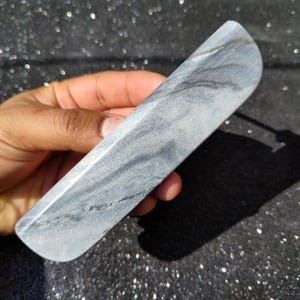 Puede incluir: Una varita de piedra gris y blanca con un extremo curvado. La varita tiene una superficie lisa y pulida con patrones de vetas naturales. La varita se sostiene en una mano, y el fondo es oscuro y brillante.