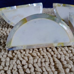 Peut inclure: Quatre sous-verres en forme de demi-cercle, de couleur blanche et irisée, avec un incrustation de nacre. Les sous-verres sont posés sur une surface en tricot beige.