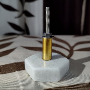 Pode incluir: Um puxador decorativo para arm&aacute;rio com base hexagonal em m&aacute;rmore branco, corpo cil&iacute;ndrico dourado e parafuso roscado prateado. O puxador &eacute; projetado para m&oacute;veis e adiciona um toque de eleg&acirc;ncia.