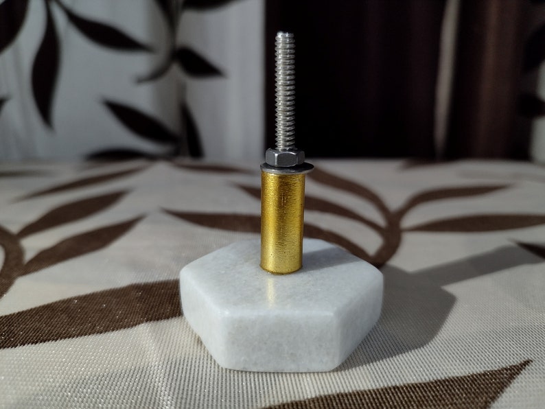 Pode incluir: Um puxador decorativo para arm&aacute;rio com base hexagonal em m&aacute;rmore branco, corpo cil&iacute;ndrico dourado e parafuso roscado prateado. O puxador &eacute; projetado para m&oacute;veis e adiciona um toque de eleg&acirc;ncia.