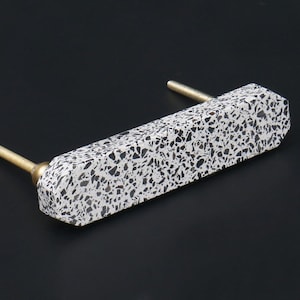 May include: A white and black terrazzo drawer pull with brass accents. The pull is rectangular in shape and has a slightly rounded edge.