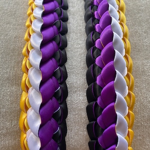 May include: Two yellow, white, purple, and black braided ribbon leis. The leis are made of satin ribbon and are braided in a traditional Hawaiian style.